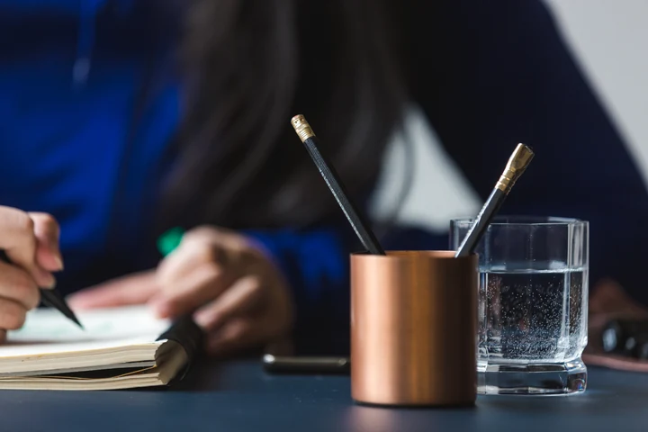 Anteckningar för hand i en öppen bok, med pennor och ett vattenglas på arbetsbordet.