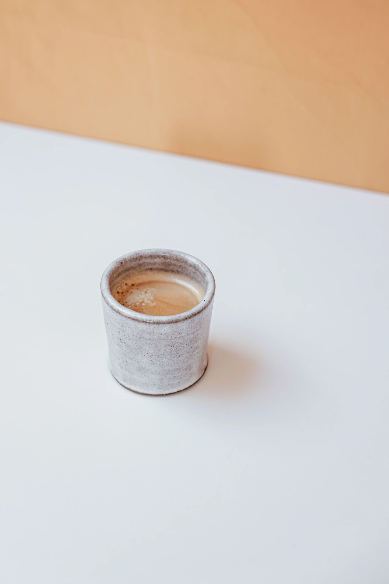 En ljus keramikkopp med kaffe, fotograferad snett från sidan mot en varm vägg.